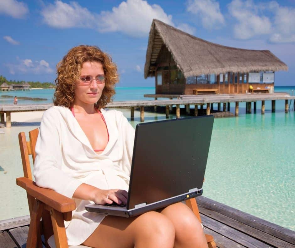 Woman with curly hair in a white cover-up sits on a wooden deck chair, typing on a laptop by a tropical beach.
