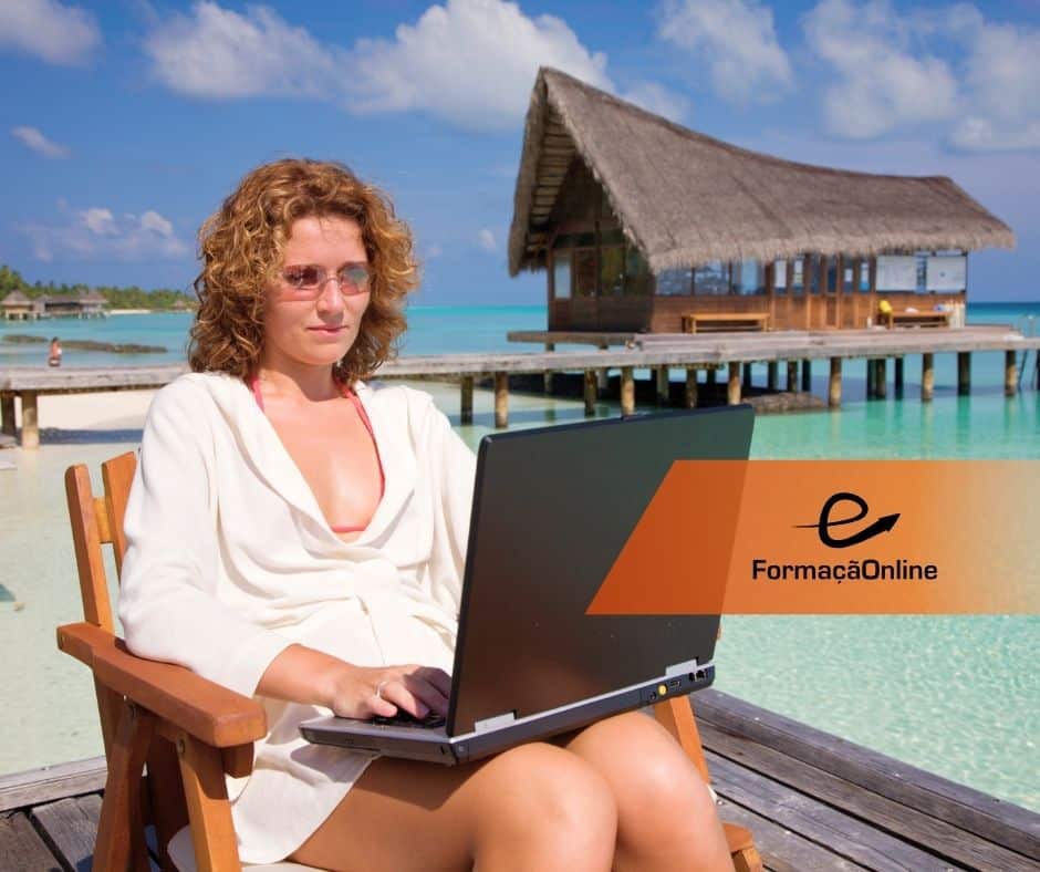 Woman in a white outfit sitting on a wooden chair, working on a laptop on a tropical beach with turquoise water and a thatched-roof bungalow in the background.