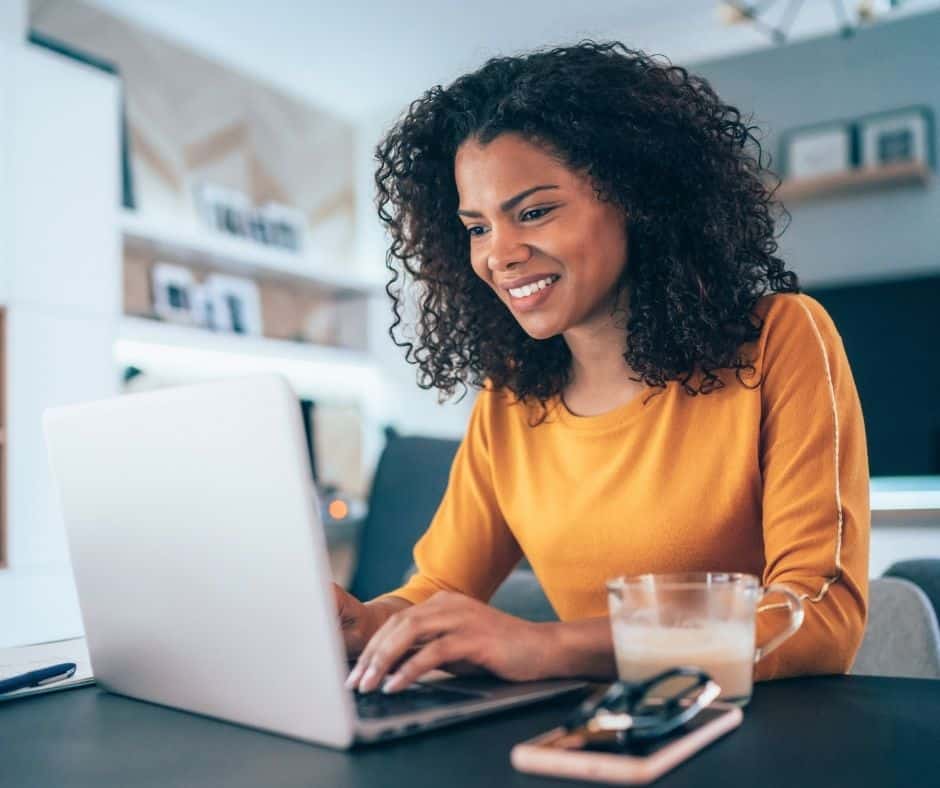 Smiling woman in an orange sweater types on a silver laptop at a desk, with a mug of coffee nearby in a bright workspace.