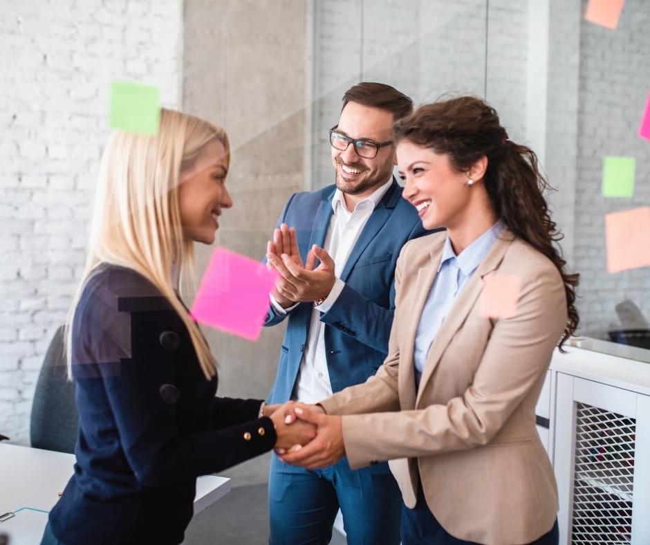 Three colleagues shake hands in a bright office, with colorful Sticky notes on a glass wall behind them.