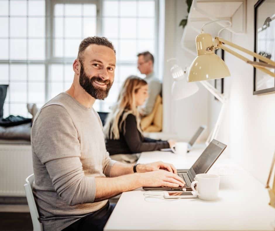 Smiling man with a beard typing on a laptop at a bright office desk, coworkers blurred in the background with a coffee cup nearby.