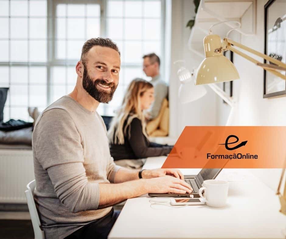 Man with a beard smiles while typing on a laptop at a bright shared workspace, with coworkers in the background and an orange FormaciónOnline banner overlay.