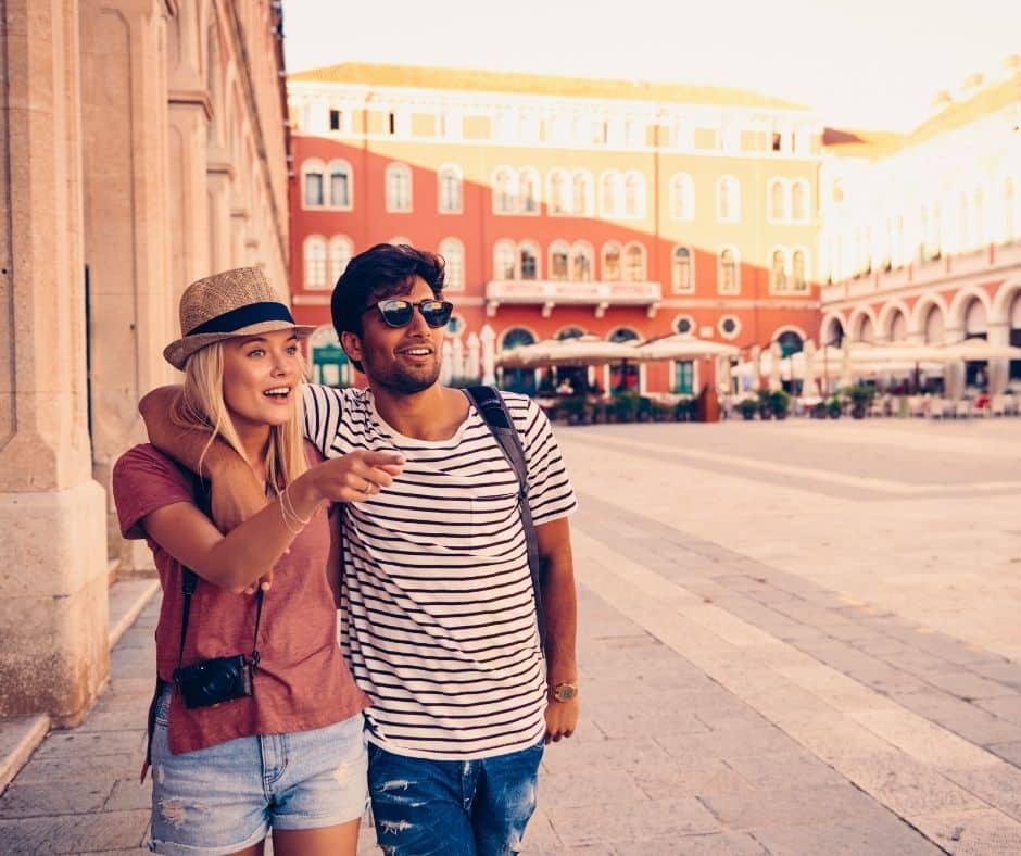 Smiling couple sightseeing in a sunny square; the woman points ahead, wearing a hat with a camera, and the man wears sunglasses and a striped shirt.