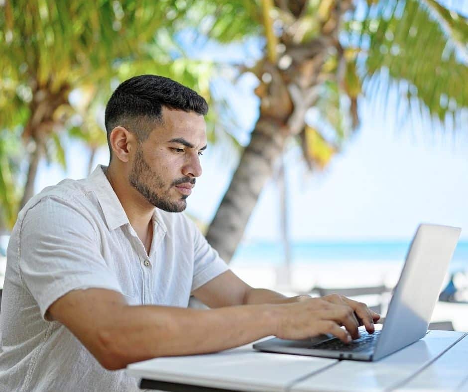 Artigo 2 Julho 2026 I | FormaçãOnline Man in a light shirt typing on a laptop at an outdoor beachside setting with palm trees behind him, sunlight filtering through leaves.