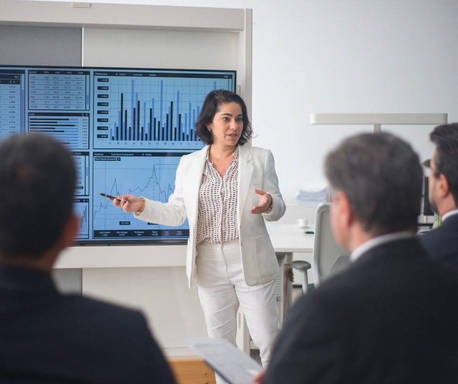 Businesswoman presenting data charts on a large screen to colleagues in a meeting in a bright office.