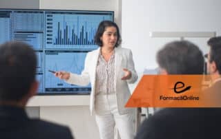 Woman in a light suit presents to a seated audience, pointing at a large screen with charts and graphs in a modern office; orange FormaçãoOnline banner overlays the foreground.