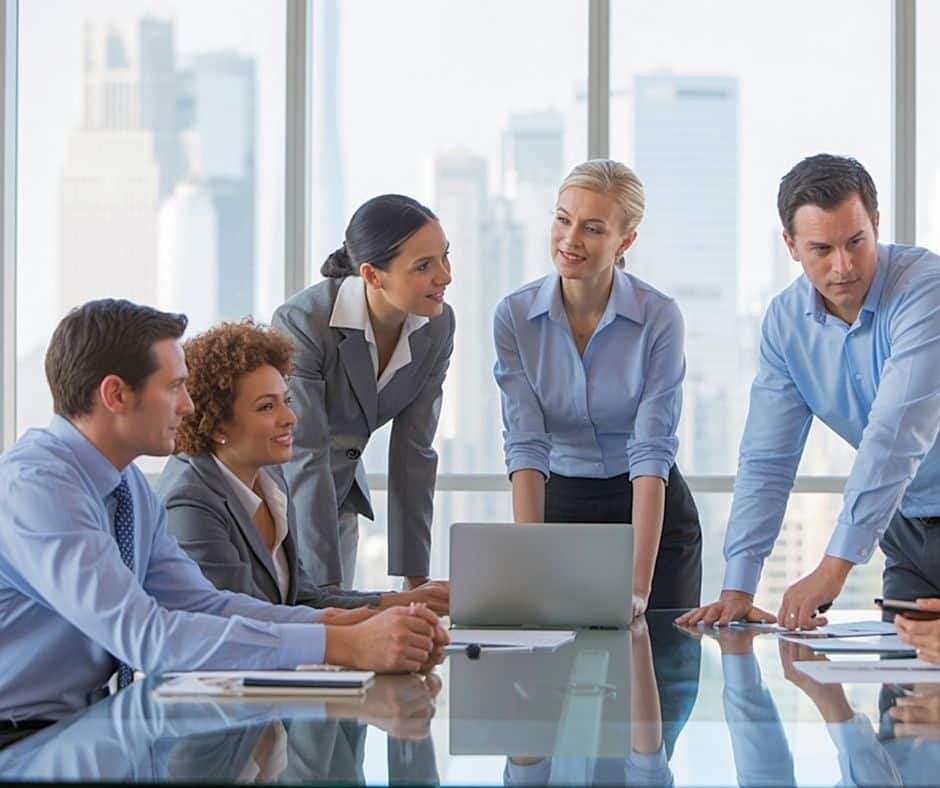 Five professionals around a glass conference table, laptop at center, in a bright office with a city skyline view wony background like blurred skyscrapers.