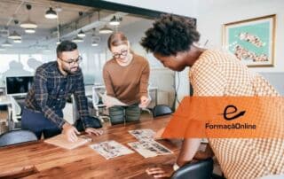 Three coworkers lean over a wooden conference table, reviewing photos and papers in a bright open office, with an orange FormaçaoOnline banner in the foreground.