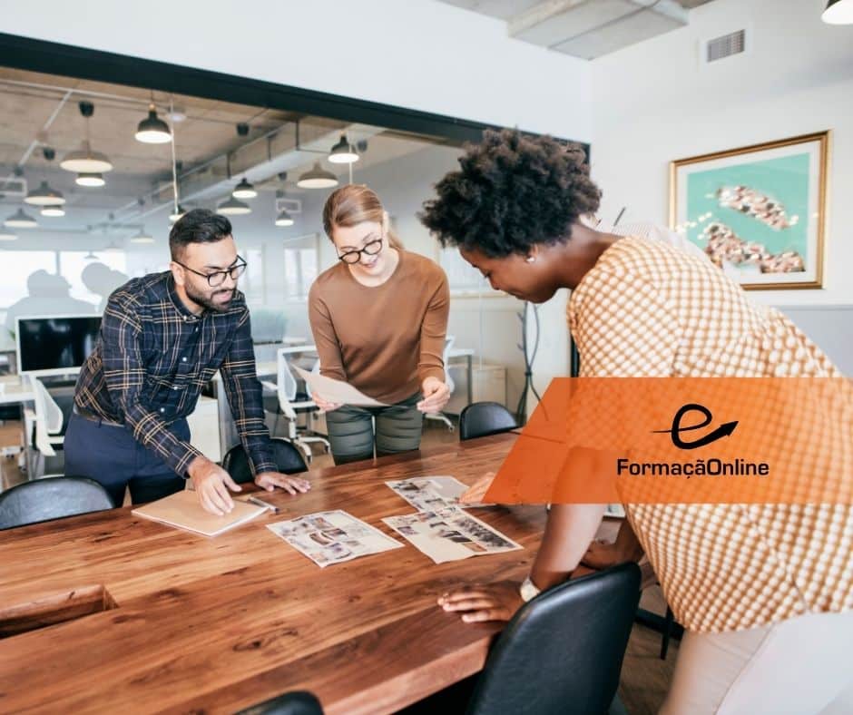Three coworkers lean over a wooden conference table, reviewing photos and papers in a bright open office, with an orange FormaçaoOnline banner in the foreground.