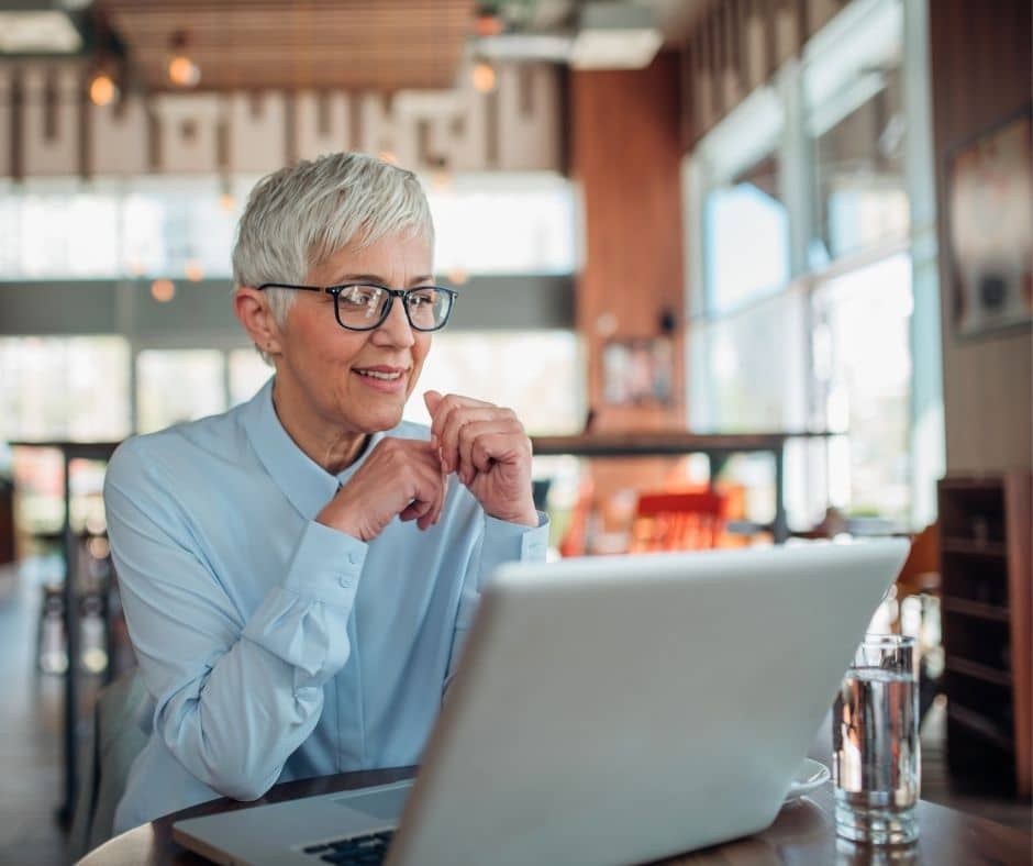 Older woman with short gray hair and glasses sits at a cafe table, looking at a laptop with a thoughtful smile.