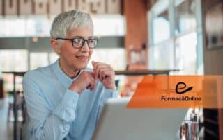 Older woman with short gray hair and glasses smiles while using a laptop in a cafe; an orange 'e FormaçãoOnline' banner is visible. (informative)