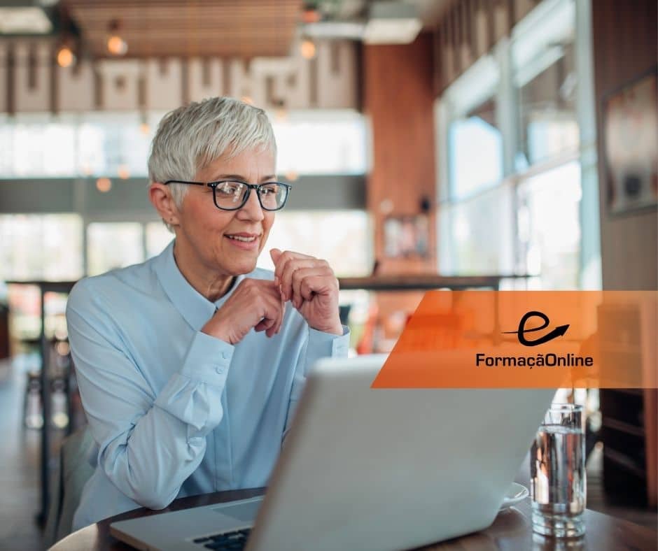 Older woman with short gray hair and glasses smiles while using a laptop in a cafe; an orange 'e FormaçãoOnline' banner is visible. (informative)