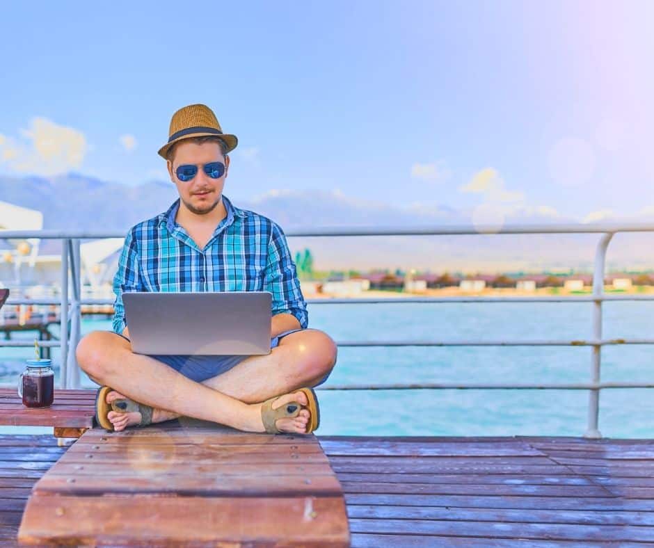 Man in a plaid shirt, hat, and sunglasses sits cross-legged on a wooden dock, using a laptop with a drink beside him by the water.