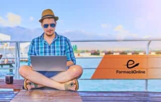 Man in a blue plaid shirt and straw hat sits cross‑legged on a wooden jetty, using a laptop by the water with a branded banner in the background ('FormaÃ§Ã£oOnline').