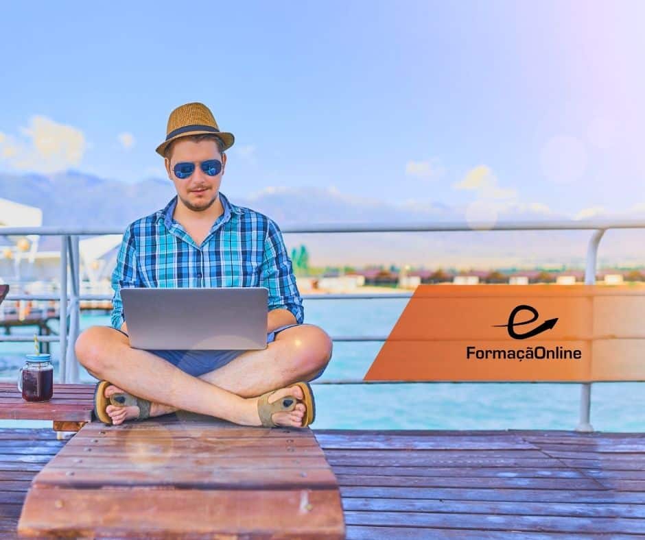 Man in a blue plaid shirt and straw hat sits cross‑legged on a wooden jetty, using a laptop by the water with a branded banner in the background ('FormaÃ§Ã£oOnline').