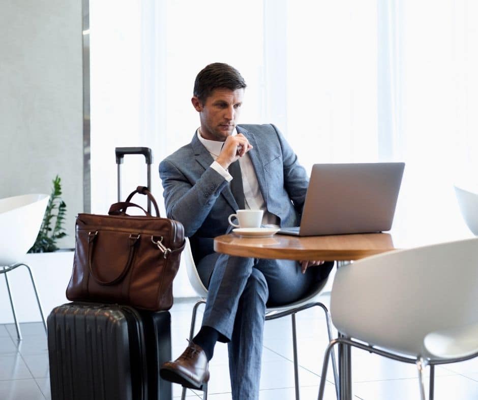 Businessman in a gray suit works on a laptop at a round wooden table in a bright lobby, with a brown briefcase and rolling suitcase beside him.