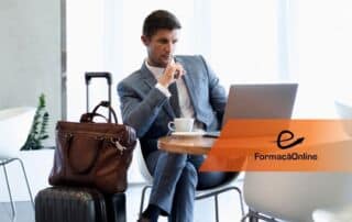 Businessman in a gray suit sits at a table with a laptop and coffee in a bright lounge; brown briefcase and rolling suitcase beside him, orange FormaçãoOnline banner across the image.