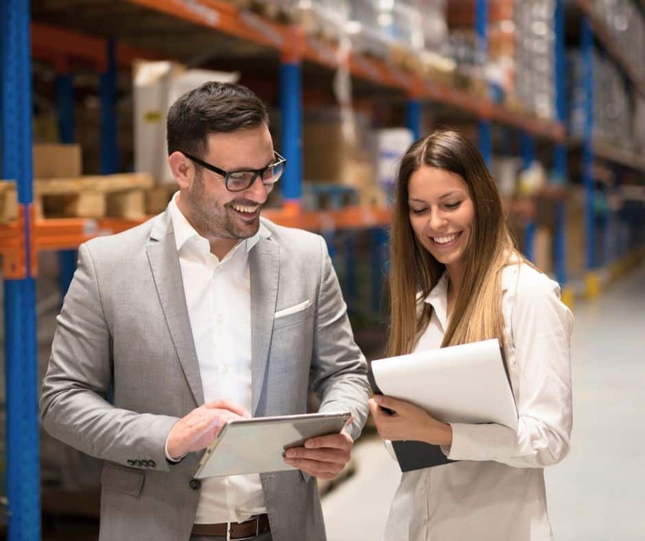 Two coworkers in a warehouse smile while reviewing information on a tablet together.