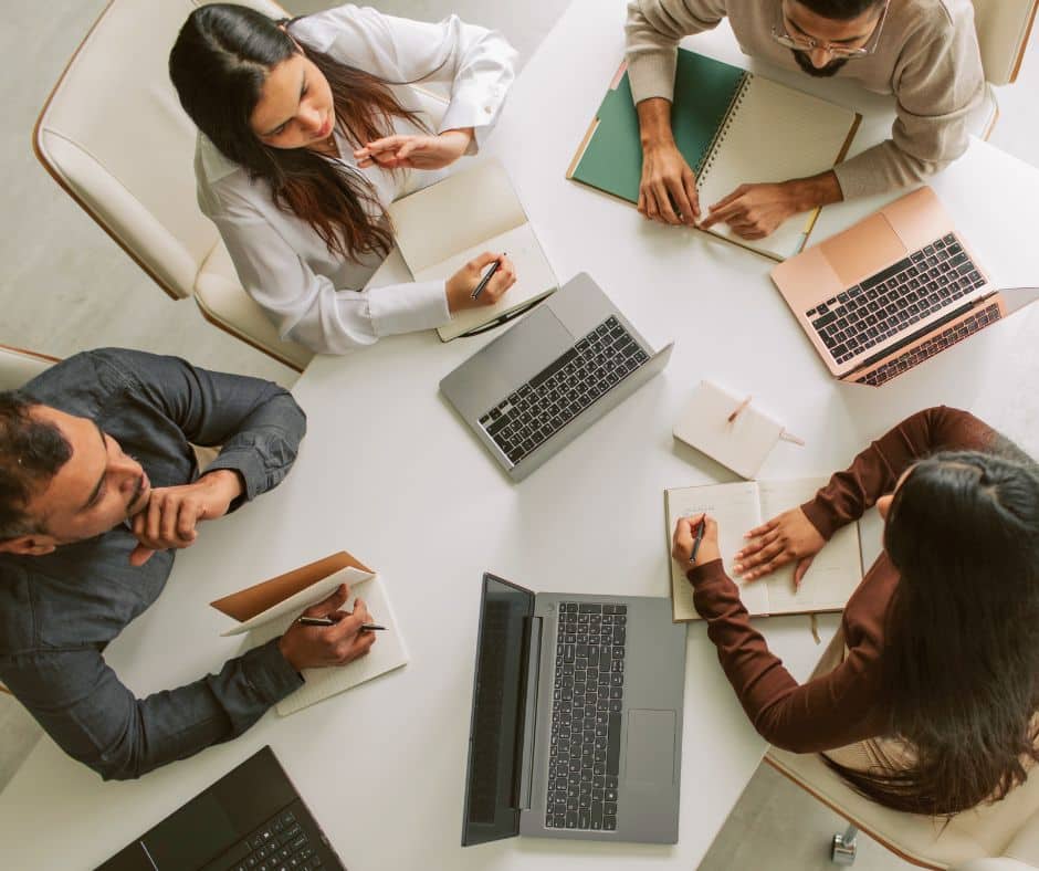 Overhead view of a team around a round white table with laptops, notebooks, and writing tools in use during a meeting.