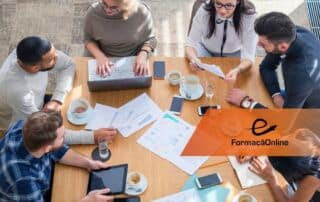 A diverse team of six working around a wooden table with laptops, papers, and coffee cups during a meeting, with an orange Formação Online logo overlay.